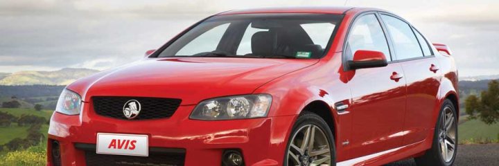 Car Hire in New Zealand - Page 1 - New Zealand - PistonHeads - The image depicts a vibrant red Avis car parked on the side of a road, showcasing its sleek design and alloy wheels. The car is set against a serene backdrop of lush greenery and a hint of mountains under a partly cloudy sky. The lighting suggests either early morning or late afternoon, providing a soft glow on the car and its surroundings. The parking spot appears to be on the side of a rural road, contributing to a peaceful and natural setting.