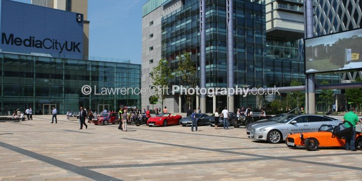 A city street filled with lots of traffic - Pistonheads - The image depicts an urban scene featuring a square with a modern architectural backdrop that includes tall buildings. The square is bustling with activity, with several people milling about. On the right side of the image, a variety of cars are parked, including one orange vehicle resembling a motorcycle or a three-wheeled car. The area appears to be a public space, possibly outside an office building, indicated by a prominent sign reading "MEDIACITY UK." The presence of numerous people and cars suggests it might be a busy day or the location popular for congregations. The image seems to be a candid snapshot, possibly for a photography website, as indicated by the text in the image.