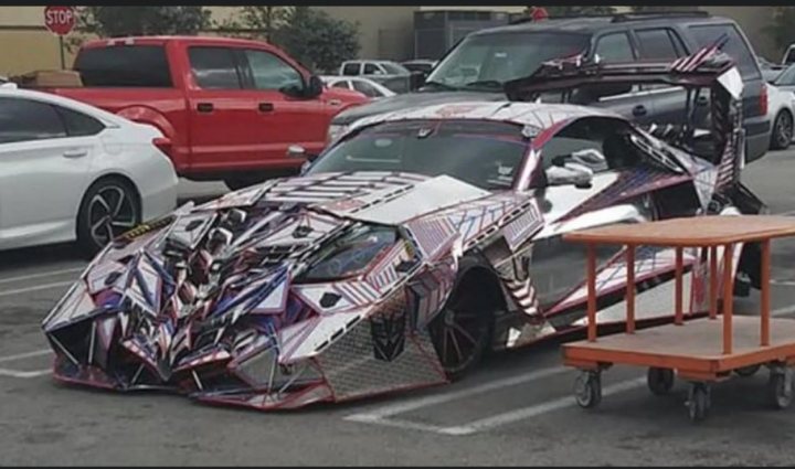 What's underneath all this? - Page 1 - General Gassing - PistonHeads - This image features a highly customized sports car parked in a parking lot. The vehicle has a unique design with a prominent front-end, including an oversized rear spoiler and side skirts. It is covered in a complex pattern of metallic shapes, possibly representing the word "Bruce" or a similar word due to its arrangement. The car also appears to have some sort of armor or protective coating on its exterior. There are multiple tables visible behind the vehicle, suggesting that this might be a public parking area. The image is in color and has a casual, candid quality.