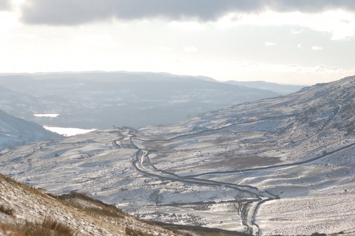 Help identify this Lake District location? - Page 1 - General Gassing - PistonHeads - A mountainous landscape blanketed in a layer of snow. A winding dirt road zigzags through the valley, surrounded by majestic snow-capped hills. The photo is taken from a high vantage point, overlooking the vastness of the snowy range. The scene is clear, suggesting it's a bright, sunny day, with the low-angle sunlight casting long shadows on the snow. The distant mountains offer a serene backdrop, conveying a sense of tranquility and solitude.