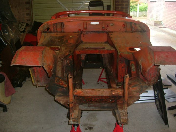 Midget Resto - Page 1 - MG - PistonHeads - The image shows a vintage car in a workspace, which appears to be a garage. The car is predominantly red, with a prominent rear wing and a spare tire mounted on its back. The vehicle has a classic design, possibly from the late 20th century. It exhibits signs of rust and wear, indicating it has seen considerable use or has been stationary for an extended period. In the background, the garage contains various tools and equipment, hinting at the location's use as a storage or repair space for automobiles.