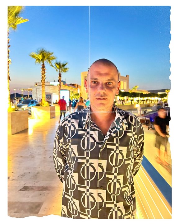 ibrahimmuratgunduz - The image shows a person standing on a walkway with palm trees, buildings, and other people in the background. The person is wearing a shirt with a unique black and white pattern. The setting appears to be a sunny day in a warm climate, possibly a tourist area.