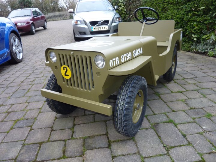 Boys Pistonheads Toy Ultimate - The image features a vintage military-style jeep, painted in a muted green color, parked on a brick driveway. The car is an antique, with a prominent number "2" on the front grille. It has an old-fashioned look, reminiscent of jeep models from the mid-20th century. There are no visible windows, and the rear-view mirror is also not present. The car has large, all-terrain tires that are currently not in contact with the ground, due to it being jacked up. There are other cars parked in the background, suggesting that the jeep is on a residential street or in a driveway. The surrounding area features a well-maintained lawn and some greenery, indicating a garden or a park.