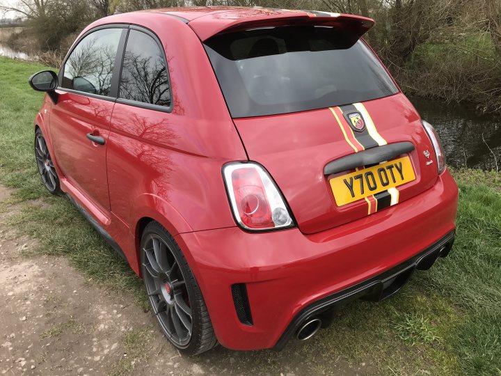 What private plates do you have? - Page 68 - General Gassing - PistonHeads UK - The image shows a red car parked on grass. It's a sports car with a visible rear spoiler, and the license plate is from the United Kingdom, indicating it might be in England. There are modifications to the vehicle, such as custom bodywork and an aftermarket exhaust system, suggesting the car has been upgraded or modified for performance purposes. The background is overcast with greenery, and the style of the photograph appears candid, possibly taken by a passerby or someone close to the car's owner.