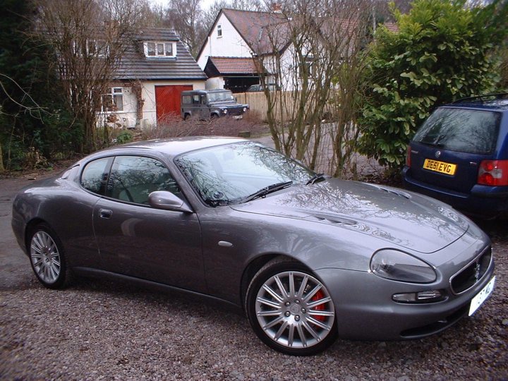 Pistonheads - The image features a gray sports car parked on a gravel driveway. The car has a sleek design with a prominent nose and alloy wheels. In the background, a blue hatchback is parked, partially obscured by a tree and plants. The setting suggests a residential area with a house and trees visible. The driveway appears to be located close to the house.