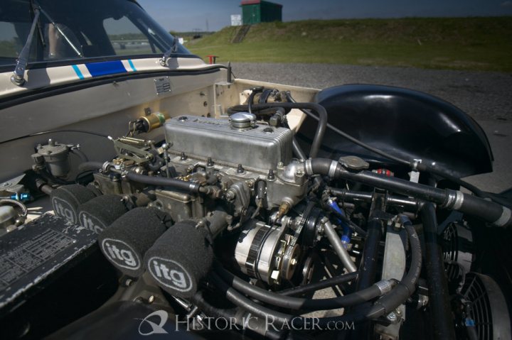 Early TVR Pictures - Page 4 - Classics - PistonHeads - The image features the engine compartment of a motorcycle, which is located in the front fairing of a classic car with an open hood. The motorcycle's engine assembly is prominently displayed with metallic parts and black components. It bears the "Itg" inscription, which could either be the brand or model name of the engine. There is also the text "HistoryRacer.com" present on the car. The scene is set outdoors with a clear sky and some foliage in the background. The engine appears detailed and well-maintained, suggesting the vehicle has been cared for and enjoyed.