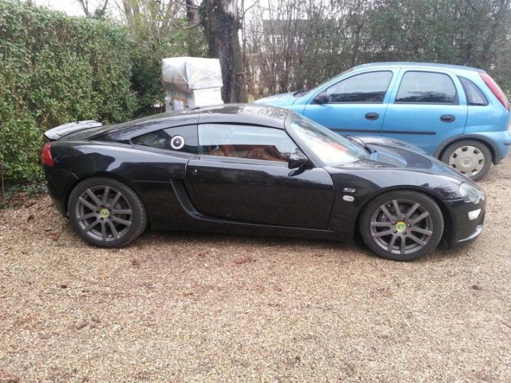 A car parked in a parking lot next to a car - Pistonheads - The image shows a driveway with two cars parked. On the left, there is a black sports car with alloy wheels, its front indicating it is a manual transmission vehicle, likely a convertible due to its sleek design and the soft top. To the right, there is a blue hatchback car with a small spare wheel mounted on the back door. The driveway appears to be a gravel road with some grass and plants along the side. The sky is overcast, suggesting it might be an overcast or cloudy day.