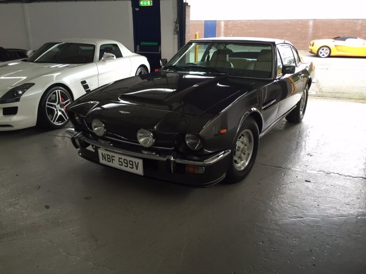 So what have you done with your Aston today? - Page 165 - Aston Martin - PistonHeads - The image shows a scene with two classic cars parked inside a building. The car in the foreground is a vintage black coupe with the text "NBF S597W" on its license plate. Behind it is a classic white convertible, and they are set against a white wall. In the background, you can see other vehicles and what appears to be a yellow exotic car. The garage setting suggests that this could be a collection or a showroom.