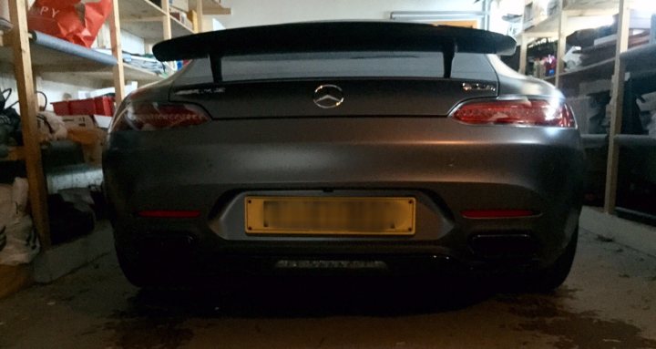 Anyone ordered an AMG GT-S yet? - Page 25 - Mercedes - PistonHeads - The image shows a sleek gray sports car parked inside a garage. The car's rear spoiler is significantly extended, giving it an aerodynamic look. The license plate is prominently visible at the back of the car. Behind the car, there is a well-organized garage storage area with shelves and baskets, indicating a neat and tidy environment. The lighting inside the garage casts a slight reflection on the car's surface. The image conveys a sense of the owner's pride in their vehicle and the careful organization of their space.