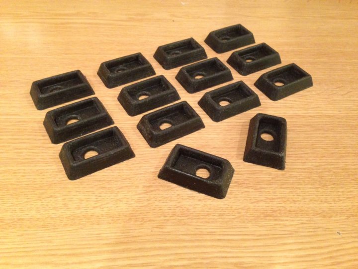 A collection of different types of remote controls - Pistonheads - This image displays a collection of eight polyhedron-shaped objects, possibly molds or containers, which are primarily black in color. They appear to be made of a somewhat rough, textured material, reminiscent of a plasti-like substance. The eight items are scattered in various positions across a wooden surface, rather than being arranged in a specific pattern or set. The lighting in the photograph casts shadows beneath the objects, indicating they are not directly sitting on the surface but rather elevated.