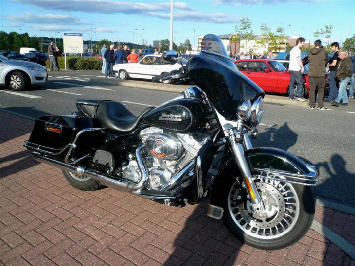 Pistonheads - The image showcases a motorcycle parked on the side of a road, facing towards the left of the frame. The motorcycle is black and has a sleek design with visible chrome details and a large engine. The setting appears to be a parking lot, as there are other cars parked in the background. People are milling around in the background, suggesting a public place or event. The scene is bathed in natural light, indicating it might be daytime.