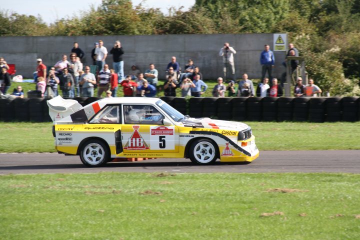 September Day Castle Pistonheads Saturday Coombe Rally - The image showcases a dynamic scene of an automobile race. A distinctive yellow and white car is speeding around a track. The number "5" is prominently displayed on the car, suggesting it might be a significant race number. The car appears to be in the foreground of the image, emphasizing its motion. In the background, there is a crowd of spectators, dressed in various styles of clothing, visible above a low chain-link fence. Their presence adds an element of excitement and anticipation to the scene. The spectators seem to be focused on the car, indicating the intensity of the race is captivating their attention. The setting appears to be a sunny day, as indicated by the clear visibility of the scene.