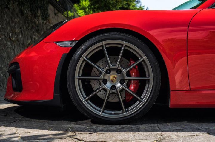 GT4 colours  - Page 52 - Boxster/Cayman - PistonHeads - This image showcases the front wheel of a vibrant red sports car, possibly a Porsche, given the distinctive hood and badge. The wheel, which is gripping an unseen surface, features a silver hub and silver spokes, contrasting with the deep red and black center. The background is blurred but suggests a driveway or a staged outdoor setting with greenery, indicating an environment conducive to a sporty experience. The focus is solely on the car, making it the main subject of the image.