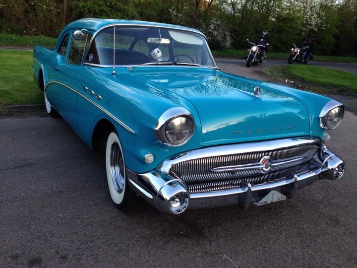 A classic car is parked in a parking lot - Pistonheads - The image shows a vintage car parked on a paved surface with two motorcycles nearby. The car is a classic model, its body painted in a vibrant shade of blue. It has a streamlined grille with the manufacturer's emblem at the center, and the chrome detailing is shiny and catches the light. The hood and the roof of the car appear to be in pristine condition. In the backdrop, you can see trees and a grassy area, indicative of a park or a similarly relaxed outdoor setting.