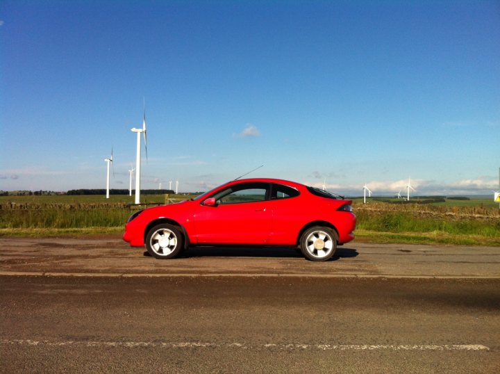 RE: Ford Puma: Catch It While You Can - Page 5 - General Gassing - PistonHeads - The image features a small red car parked on the side of a road. The car is positioned on the inside of the road, near the shoulder. Behind the car, there is a picturesque rural scene with rows of wind turbines stretching into the distance. The landscape is lush and green, and the sky is clear and blue. A single white sign with black lettering is visible in the distance, although the text is not legible in the image. The overall setting suggests a road trip or a visit to a region with a significant focus on renewable energy.