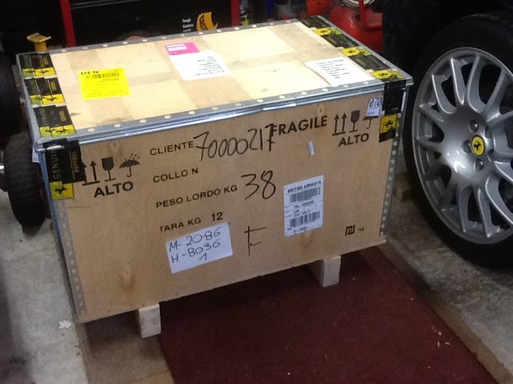 Merry Xmas to me .. 360 content - Page 1 - Ferrari V8 - PistonHeads - The image shows a cardboard box with fragile items intact and properly labeled. The box has the text "CLIENTE 000021" and "ALTO" suggesting it is fragile and should be handled with care. There is also text that reads "ATTORGERIA" and "peso loro ko 38," possibly indicating the item's origin or purpose. The numbers "M 2086" and "H 01" might be related to an inventory or shipping system. The box is placed on a floor inside what appears to be a building or garage, as a shelf and part of a vehicle can be seen around it.