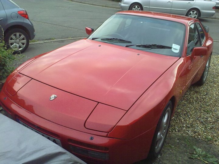 Exist Didnt Pistonheads Mxs - The image shows a bright red sports car parked on the side of a street next to a gray vehicle. The sports car has a sleek design, with a visible hood emblem, suggesting it's a Porsche. The car has a convertible roof, giving it a distinctive and luxurious look. There is a person partly visible within the sports car, possibly the driver. The overall setting appears to be a city street.