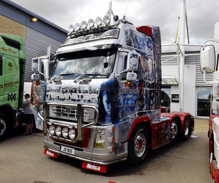 Truckers and Airbrushing  - Page 1 - Commercial Break - PistonHeads - The image showcases a large, silver semi-truck parked on a street. The truck is prominently customized with a vibrant, blue and red digital painting superimposed on its exterior, transforming it into a moving work of art. This striking image takes up the majority of the frame, emphasizing its size and the intricate detailing of the artwork. In the background, another truck can be seen, indicating a commercial or industrial setting. The presence of numerous people in the vicinity, along with a building and a railing, provides context for the vehicles' location.