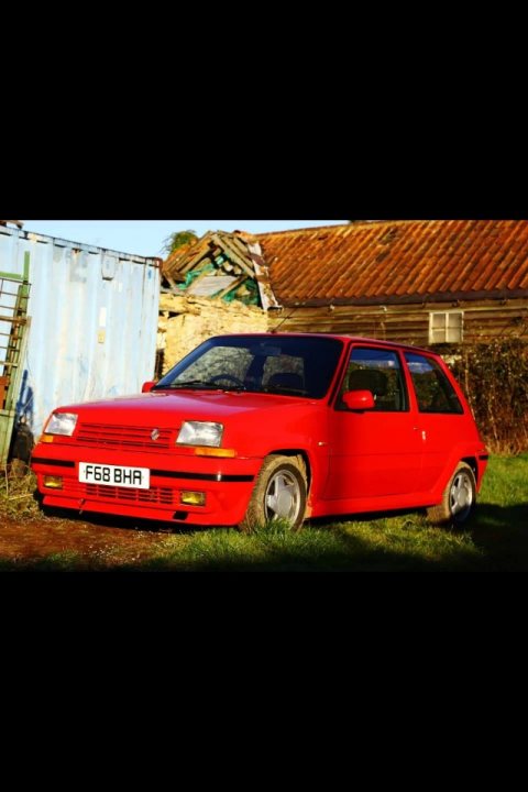 Renault 5 GT Turbo Wanted - Page 1 - French Bred - PistonHeads - The image is a photograph showing a red hatchback car parked on a grassy area in front of a rusted metal building. The sky is clear, suggesting it might be late afternoon or evening. There are no people or moving objects visible in the scene, giving a somewhat still and quiet atmosphere. The fallen tiles of the roof next to the car indicate potential neglect or abandonment of the structure.