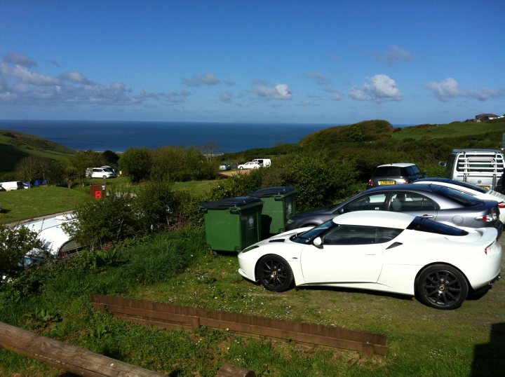 Random Evora Pr0n... - Page 1 - Evora - PistonHeads - The image captures a serene coastal scene. Several cars are parked on either side of the frame, with one prominent white sports car taking the center stage. The cars are parked on what appears to be a grassy area, possibly a parking lot or a long driveway, with a fence or hedge dividing the space. In the background, there's a vast expanse of water under a clear sky, with clouds indicating a sunny day. This coastal location adds to the overall tranquility of the scene.