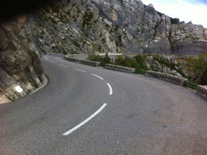 Driving France - Page 4 - Chimaera - PistonHeads - The image captures a serene scene on a winding asphalt road. The road, bordered on one side by a low stone barrier, cuts through a mountainous region. The road's white boundary lines contrast starkly against the gray of the pavement, guiding the way for vehicles traveling around the bends. On the right side of the photo, the rocky terrain of the mountains is partially encroached upon by wild, overgrown vegetation. There's no visible traffic in the image, adding to the tranquil atmosphere of the scene.