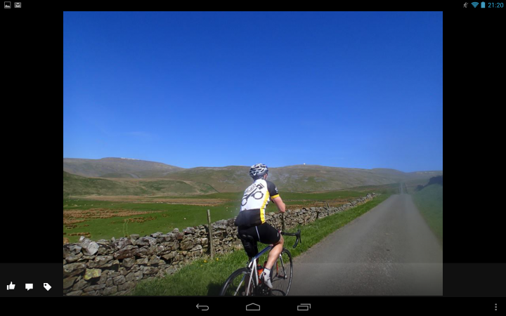 A man riding a bike down a dirt road - Pistonheads - The image captures a person riding a bicycle on a paved road. The road winds through a sunlit landscape, with a stone boundary and lush grass on either side. In the distance, rolling hills rise against a clear blue sky. The cyclist appears to be dressed in athletic attire, suggesting they might be on a long-distance ride or training for a race. This is a vibrant and energetic scene, filled with the joy of outdoor activity.