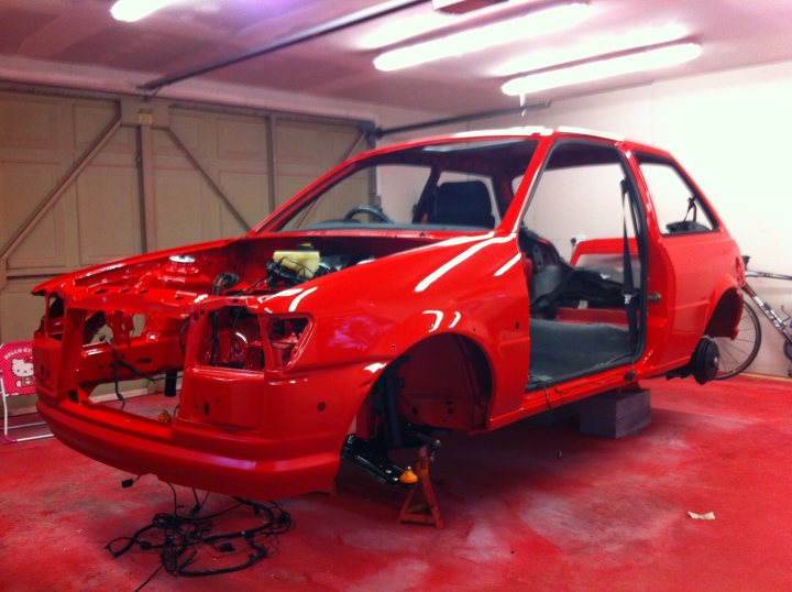 Pistonheads - The image depicts a red car in the midst of a paint job, with the hood and front portion carefully stripped, revealing the underlying engine and components. The car is depicted lying on its side with a ramp underneath for access to the work area. Various tools can be seen around the vehicle, indicating ongoing work on the car's exterior. The setting appears to be a garage or workshop, and the lighting suggests a professional environment.