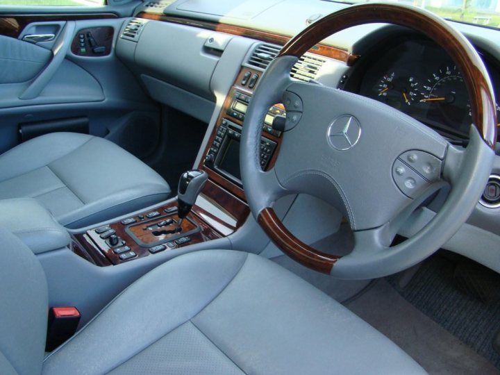 Mercedes w210 E430 (no titivating allowed) - Page 1 - Readers' Cars - PistonHeads - The image shows the interior of a Mercedes-Benz vehicle, featuring the steering wheel, dashboard, and a portion of the car's front seat. The steering wheel is prominently displayed, with theMercedes-Benz emblem at the center. The dashboard has various control knobs and buttons, indicative of the car's features. The interior of the car is in a light color, likely beige, which complements the wooden trim seen around the instrument panel and the steering wheel. The car appears to be well-maintained and is possibly parked inside a garage or under a large awning, as indicated by the reflection and light distribution visible in the image.