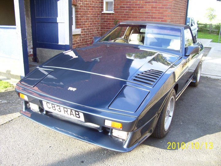 Big Pistonheads Yorkshire Sunday October Breakfast - The image features a metallic dark blue convertible sports car parked in front of a brick building. The car's top is down, revealing a sleek interior. The vehicle is adorned with a European-style license plate and has a distinctive design with a long front hood. The setting appears to be a quiet street, with the time and date "2010/10/13" overlaid on the image.