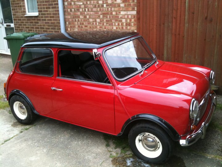 Fancy selling me your mini - Page 1 - Classic Minis - PistonHeads - The image shows a classic Mini Cooper car parked on a driveway. The car is a vibrant red with a contrasting white roof that matches the walls of a nearby house. It appears to be a vintage model, sitting in front of a brick wall and next to a green trash can. The vehicle's round white headlights and red tail lights suggest a design characteristic of the Mini Cooper brand. The scene gives off a nostalgic vibe, reminiscent of the architecture and automotive styles of the past.