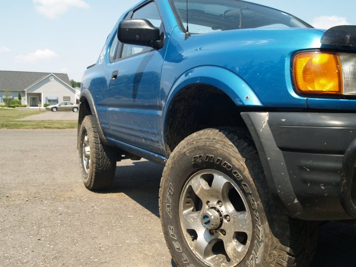 Mitsubishi Pinin lift option? - Page 1 - Off Road - PistonHeads - This image captures a scene of an old, blue pickup truck parked on a paved driveway. The truck stands out with a mud-covered front right tire, suggesting it might have been driving on an unpaved road or area. A white house with a gray roof is visible in the background, indicating a residential area. The truck's side, visible in the shot, shows signs of use and weathering, with chipped paint and scratches on the body.