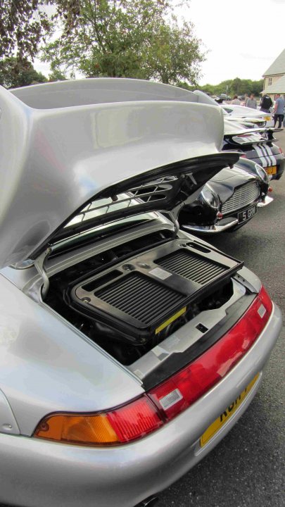 Hare Breakfast Meet - August - Page 1 - Kent & Essex - PistonHeads - The image shows the rear view of a white sports car with its hatchback trunk open. The inside of the trunk is visible, showcasing a black mat with grid lines. The car is parked among several other cars, suggesting the setting might be a car show or a crowded parking lot. The focus is predominantly on the white car, highlighting its design and the open trunk revealing the storage area. The brand of the car suggests it is a luxury or high-end sports car.