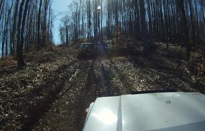 Bit of off-roading, and a bit of camping. - Page 1 - Off Road - PistonHeads - The image is a photograph from the perspective of an off-road vehicle at the rear, capturing its rearview on a dirt road. A utility task vehicle (UTV) is seen driving up a steep and uneven dirt hill. The photo is taken from inside a vehicle, with the road appearing narrow and bordered by tall brush and dense tree foliage on both sides, creating an inviting atmosphere during the day. The sky is clear, implying favorable weather conditions at the time.