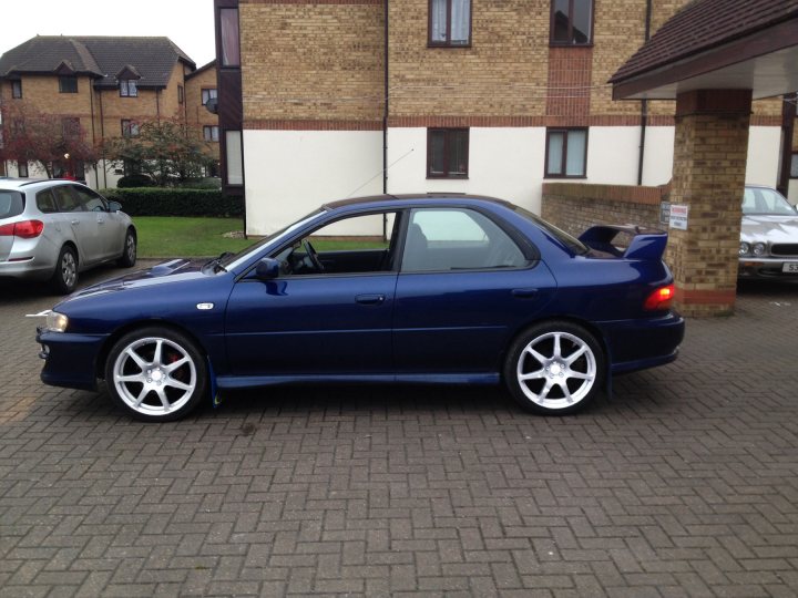 Can someone please photoshop my impreza. No idea how to :/ - Page 1 - Subaru - PistonHeads - The image shows a dark-colored, mid-size sports coupe parked on a gray brick surface. The car features a sunroof, side demister vents, and large five-spoke wheels. It appears to be a two-door model, possibly a sportsback, and has a sleek and modern design. The setting is an urban area, with buildings visible in the background and other vehicles parked nearby.