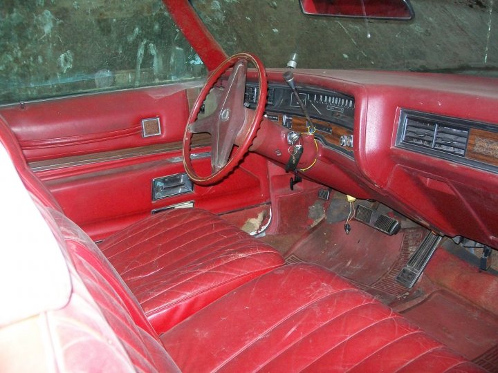 Uncle Shed Pistonheads - Pre-1980s red car interior shows worn leather seats, cracked dashboard, missing steering wheel cover, and a few dials and switches. The windows reveal a dimly lit, possibly underground, room in an older building. Door panels are missing, and a handrail has been added for support.