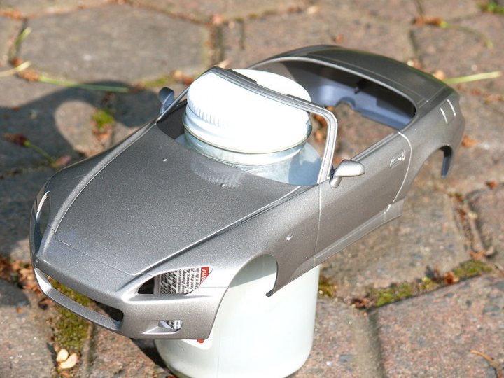 Pistonheads Honda Tamiya - The image shows a close-up of a gray plastic or toy car, which resembles a sedan model. The car is predominantly gray, with a visible darker intake vent just before the front wheels. On the hood, there's a bottle-like object attached or placed, making it appear as if the bottle is the windshield. This appears to be a playful and creative setup, likely intended to mimic a bottle of ketchup or something similar. The background is a nondescript, dark surface that looks like a stone or concrete ground.