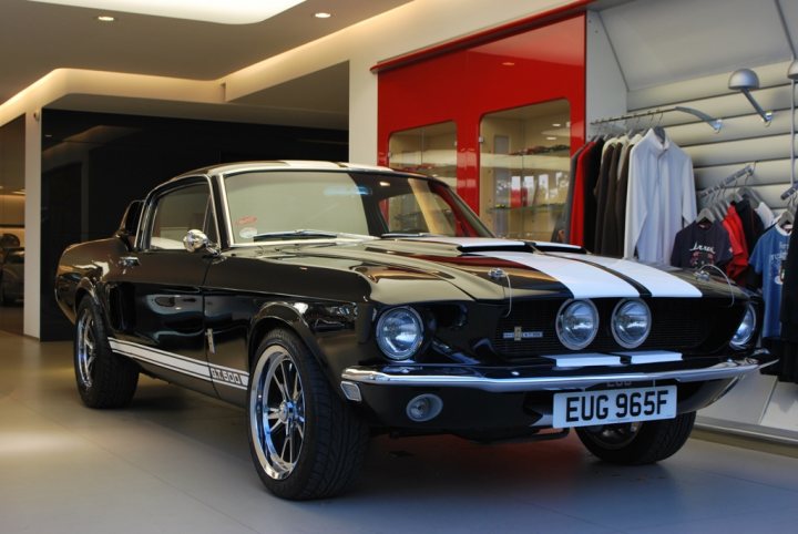 A car parked in a parking lot next to a car - Pistonheads - The image captures a striking scene in a store or showroom. The central focus is a classic black and white Ford Mustang, its sleek body and chrome detailing gleaming against a pristine gray floor. The shop is well-lit, and the Mustang is parked right next to a vibrant red door, creating a striking contrast between old and new, black and white, and red. The showroom itself is minimalist and clean, with white walls adding to the overall presentation. The Mustang's position and the absence of other cars suggest it is being showcased for sale or display.