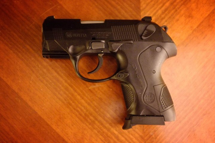 A close up of a cell phone on a table - The image features a realistic-looking airsoft gun with a matte black finish. Various textures are visible, with ridges running through the grip and along the magazines and barrel. The gun is positioned at a diagonal angle on a wooden surface, showcasing the details of its design, including the sleek slide and the weighty barrel. No additional context, such as a person holding the gun or a specific setting, is provided in the image. The focus is solely on the airsoft weapon itself.