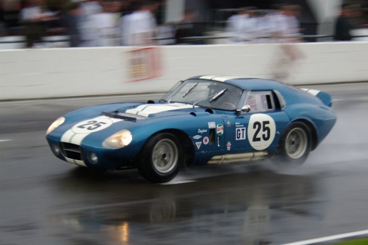 Revival Photos - Page 2 - Goodwood Events - PistonHeads - This image captures a dynamic scene on a racetrack. The central focus is a blue and white racing car, identified by the number 25 on its side. The tires of the car are blurred, suggesting that it is moving at high speed, possibly reaching 250 miles per hour, as indicated by the numbers 250mph. The background features a blurred scene of a race at night, with spectators visible. The rain on the track adds a dramatic effect to the image, creating an atmospheric distortion around the car. The image conveys the thrill and excitement of racing.