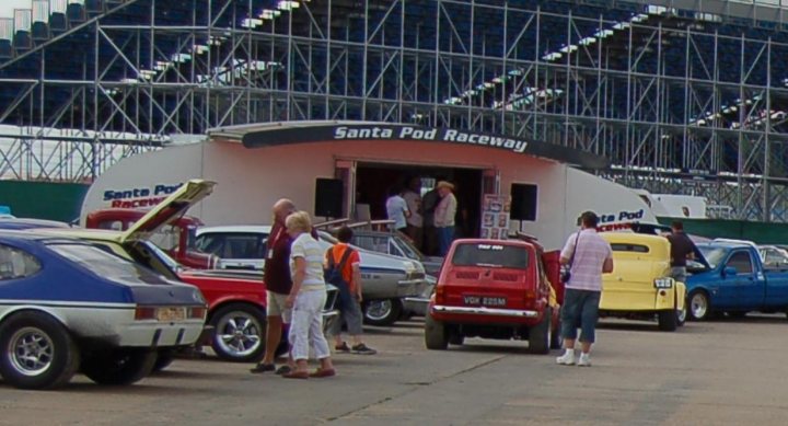 Silverstone Classic Pistonheads Goto - The image captures a vibrant scene at a racing event. A white building with the sign "Santa Pod Raceway" stands prominently in the background, surrounded by various vehicles and people. There are cars in various colors, including an unexpected red three-wheeler. People can be seen milling around, some standing in front of the white building, perhaps discussing the raceway or the day's activities. The atmosphere is busy and filled with anticipation, characteristic of a racing event.