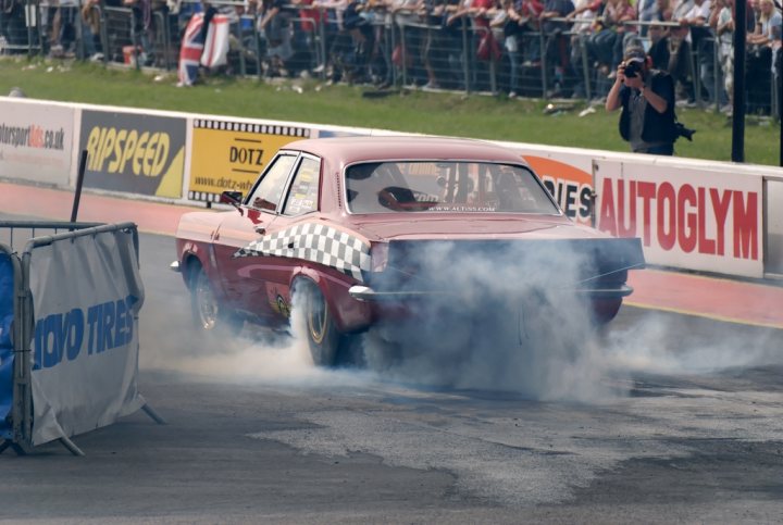 Fast Red Pistonheads Victor History Vauxhall - The image captures a thrilling moment at a car racing event. A red racing car, with a black-and-white checkered design on its hood, is seen in the foreground, emitting a billowing smoke suggesting high speed as it takes a turn on the blacktop of the track. The car is leaving the dirt-covered backstretch, as indicated by the surrounding environment. The setting appears to be a professional race track with multiple spectators in the background, adding to the excitement of the scene. The focus is on the car and the action around it, while the rest of the image is filled with the cheering crowd and the lush greenery around the track.