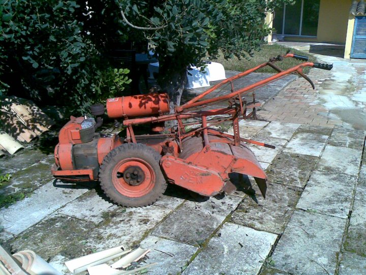 A bit of a rant!  - Page 1 - Spain - PistonHeads - In the image, there's a rusty orange tractor with large front tires and a chisel attachment facing downwards. The tractor is parked on a tiled walkway, with a side of concrete and grass visible in the background. It's near a tree and a building, suggesting an outdoor or semi-urban environment. The overall scene is somewhat worn, possibly due to the outdoor storage and the age of the tractor.