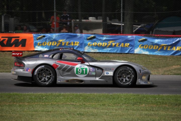 New GTS-R race debut - Page 1 - Vipers - PistonHeads - This is a color photograph of a sports car in action. The car is prominently featured in the foreground, adorned with racing number "91" and sponsored by a variety of companies such as Goodyear and Continental, among others. It is captured on a track marked with various racing indicators like kim green and mexican green. The track is bordered by spectator seating on the side, where a few people can be seen observing the race. The sky is overcast, and the grass around the track is well-maintained, indicating a well-organized racing event.