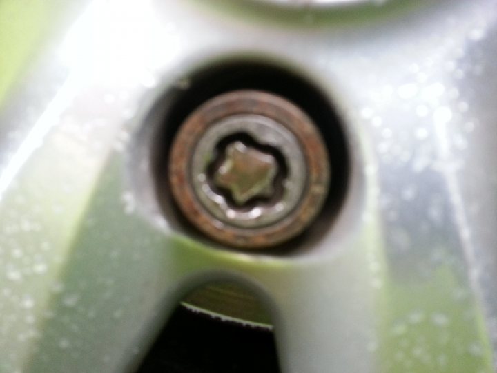 Rounded locking wheel nut removal? - Page 1 - Home Mechanics - PistonHeads - The image captures a close-up view of a car's mechanical component, possibly a wheel or brake part, with a hexagonal bolting nut at the center, surrounded by a flange. Water droplets are present on the object, partially obscuring its details.