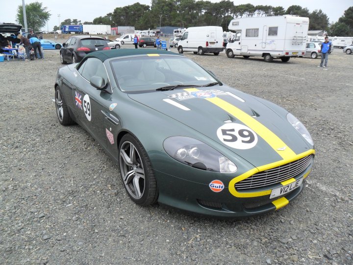 Aston Cars stickered up for LeMans. - Page 1 - Aston Martin - PistonHeads - In the heart of a gravel parking lot, a bespoke green Aston Martin sports car with white racing stripes takes center stage. The license plate "V12LJE" adorns its front, hinting at its past or future adventures, and it proudly displays "59." The environment around it is buzzing with activity, with several trucks, trailers, and other vehicles parked in the vicinity, indicating that a large event or gathering might be coming up. In the distance, a solitary figure can be seen, perhaps preparing for or returning from the day's activities.