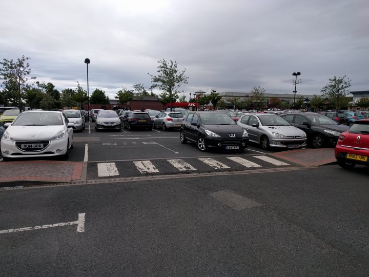 The BAD PARKING thread [vol4] - Page 3 - General Gassing - PistonHeads - The image depicts a bustling parking lot at what appears to be a shopping center. There are multiple cars parked in neat rows, facing the same direction. The main focus of the image is a black SUV parked in a space with its headlights on. The lot is filled with various models and colors of cars, indicating a diverse group of shoppers. The sky above the lot is overcast, indicating perhaps an earlier or later shopping time, suggesting this could be a common scene in this location.