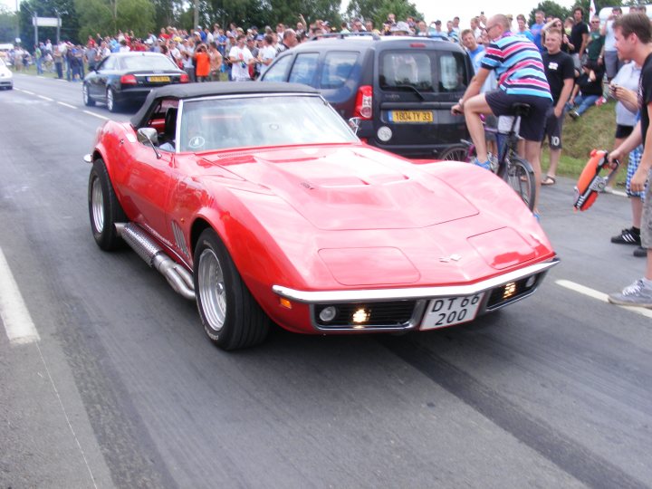 Pistonheads Classics - The image captures a vibrant street scene dominated by a striking red Corvette sports car driving down the road. The car is a convertible model, characterized by its sleek design and shiny, well-polished exterior. In the background, there are spectators lining the street, their attention focused on the Corvette, suggesting a possible car show or parade event. Various other cars and a bicycle are present on the road, adding to the sense of movement and activity. The overall atmosphere feels both dynamic and festive, with the vintage sports car as the star of the show.