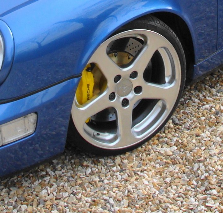 Pistonheads - The image shows a close-up view of the front tire of a blue luxury car. The tire has a silver rim with a shiny appearance and a yellow speed sensor visible on the side. The car is parked on what appears to be an asphalt or light-colored gravel surface, with the front corner of the car in focus against a softer background. The tire has eight petal-shaped spokes, common to modern wheel designs, and the brake disc is visible behind the rim. There is no text present in the image. The lighting suggests daytime with clear visibility.