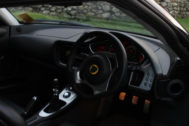 black evora spotted - Page 1 - Evora - PistonHeads - The image captures the interior of a car, focusing on the driver's side. The dash includes multiple controls and displays, including a digital panel that may be the instrument cluster. The steering wheel is black with a distinctive gold circular symbol, suggesting it's a branded item, potentially for a luxury or high-performance vehicle. The car appears to be a sports or high-end sedan given the design elements visible in the photo. It's worth mentioning that the lighting inside and outside the vehicle is dim, which might be a deliberate choice for artistic effect.