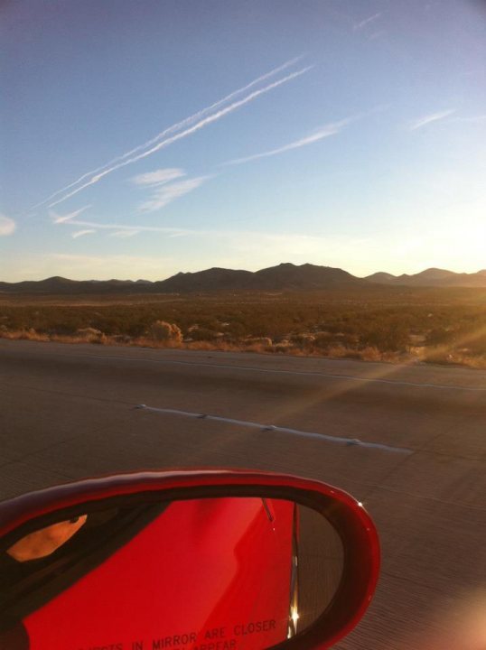 Speed cameras LA to Vegas - Page 1 - USA & Canada - PistonHeads - The image captures a vast desert landscape at sunset, as seen through the rearview mirror of a vehicle. The clear red of the rearview mirror contrasts with the open sky above, which transitions from a warm orange hue near the sunset to a lighter gradient as it reaches the horizon. The pole for the rearview mirror extends from its central position slightly to the left of the frame, providing a diagonally framed reflection of the rearview mirror and the desert scene. The stars in the sky are faintly visible against the descending sun.